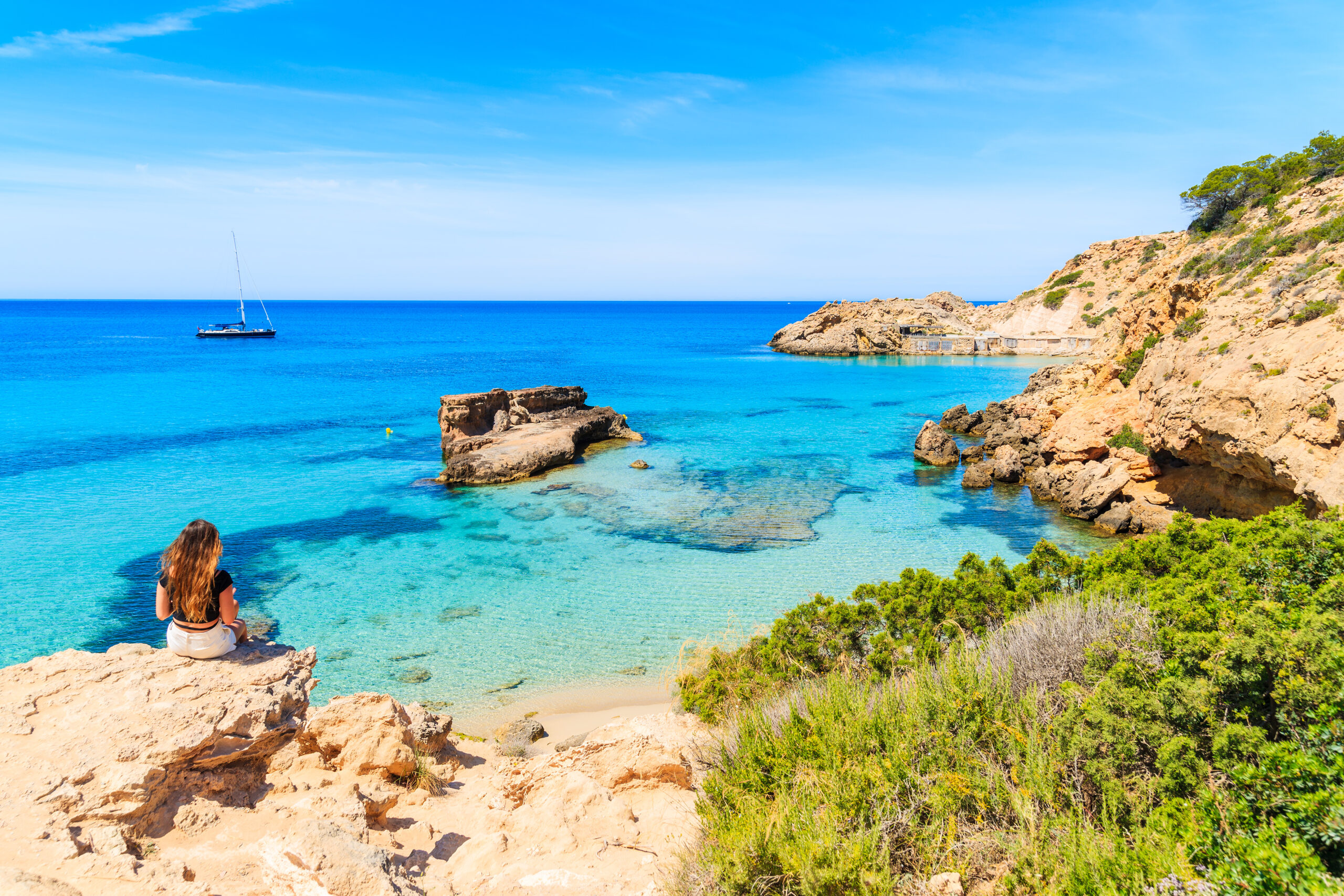 Unidentified young woman sitting on a rock and looking at beautiful Cala Tarida bay, Ibiza island, Spain